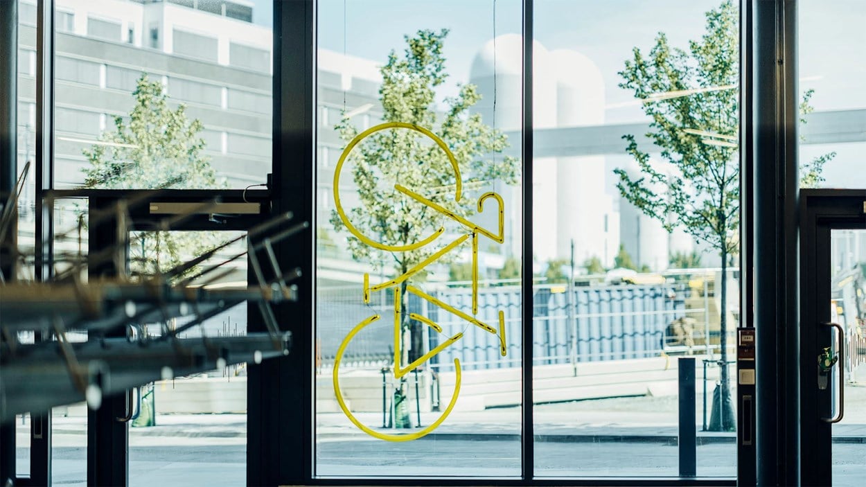 Vy innifrån ett cykelhotell med stora fönster utåg gatan. I ett av fönstren hänger en modell av cykel helt i gult.