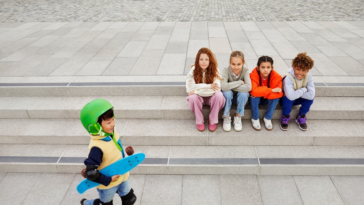 Barn som sitter på en trappa där en liten pojke går förbi med en skatenboard under armen.