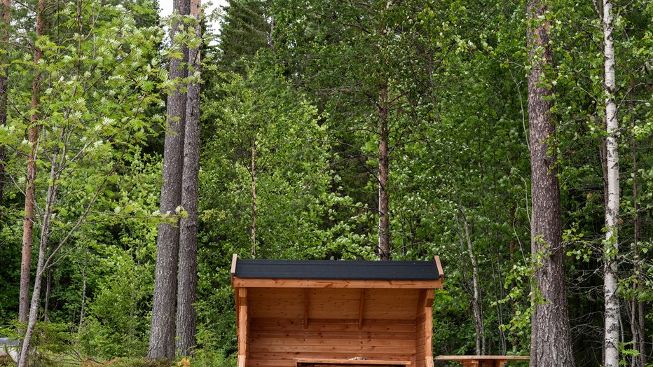 Plats för lek och utflykter nära naturen. Trähus med öppen framsida, intill finns träbord och bänkar av stockar.