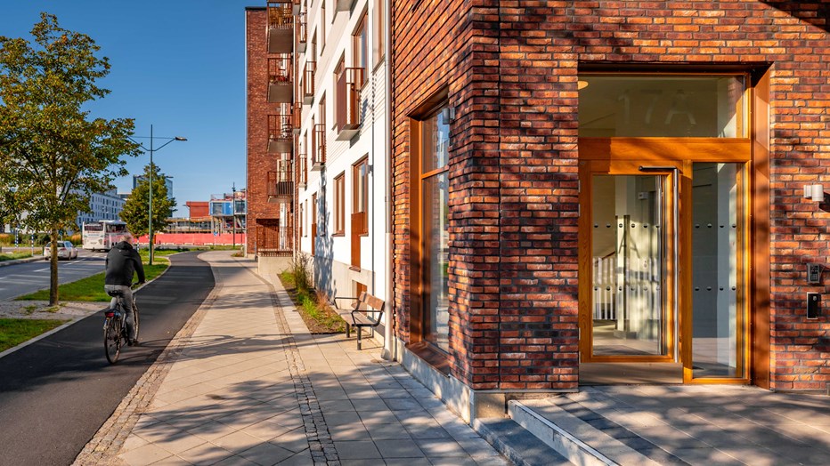 Fasader i tegel och entrépartier i oljad ek. Närbild av en hörna av fastigheten med fasad i rött tegel och portar i brunt ekträ. Runt huset finns en trottoar.