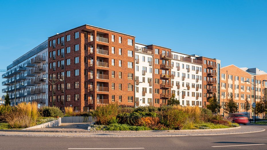 Hörnan på flerbostadshuset med vit och röd fasad. Träd och gräs nedanför och framför finns en stor gata.