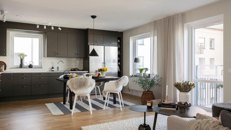 Kvarteret Scilla blir ett netto-nollenergihus vilket innebär att fastigheten har betydligt lägre energibehov än BBR-standard och tillför lika mycket förnybar energi som fastigheten behöver under ett år Kök och vardagsrum i öppen planlösning med matbord, stora fönster och soffdel.