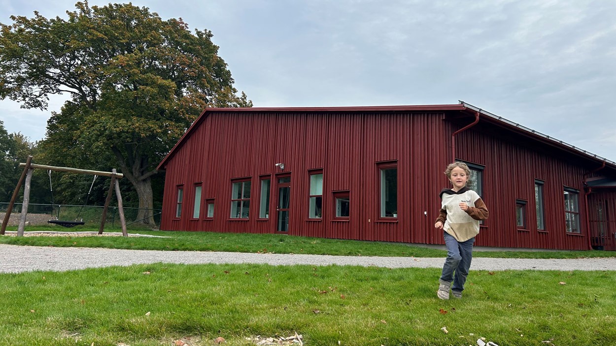 Pojke som springer i gräset framför en röd skolbyggnad med träfasad. Till vänster om skolan står en gungställning.