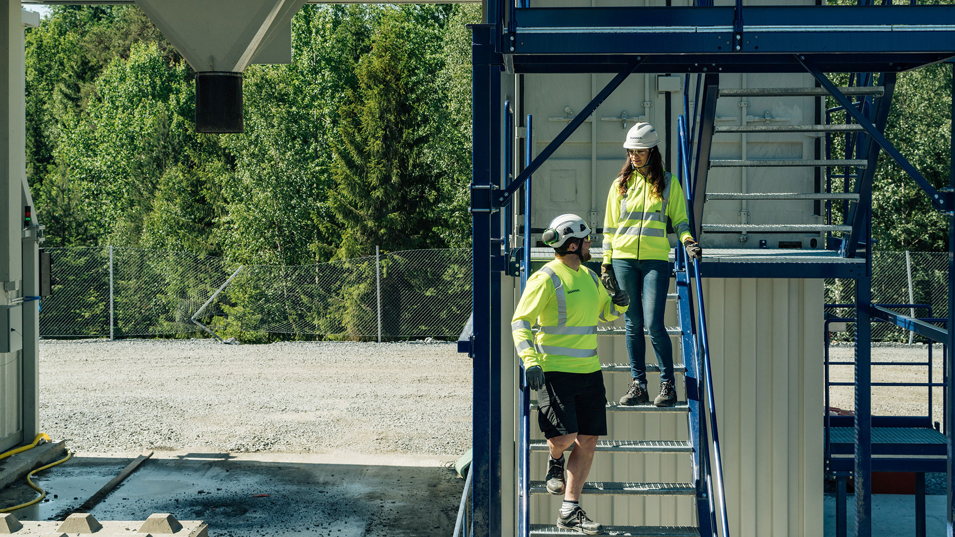 Två personer med hjälm och skyddsväst som går i en trappa utomhus.