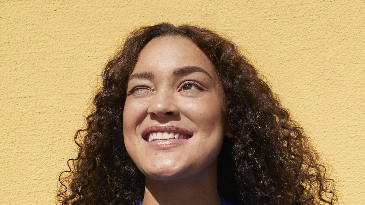 Ung kvinna som står mot en gul vägg och ler mot solen och kisar.