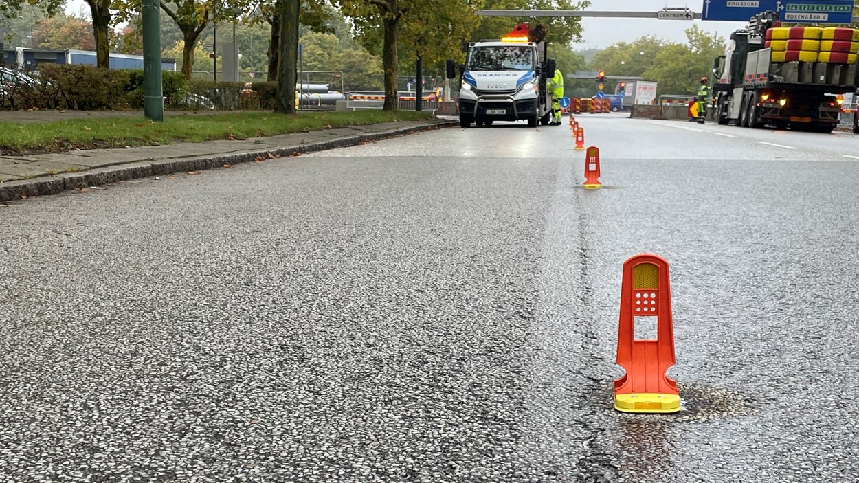 Asfaltväg med små orange fastmonterade markeringar i mitten av vägen. I bakgrunden syns fler avspärrningar och personer i varselkläder.
