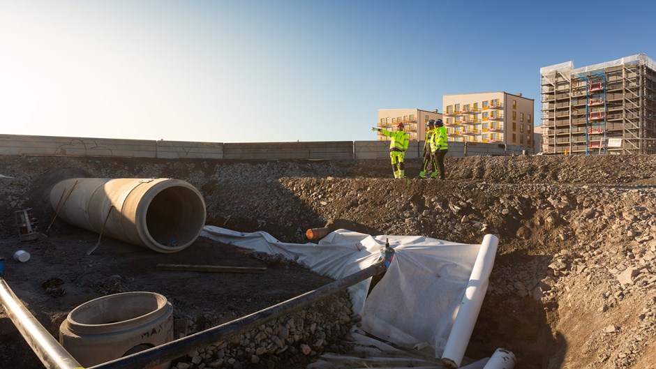 Betongrör som ligger i djupt och brett dike. Uppe på kanten står två personer i gula varselkläder.