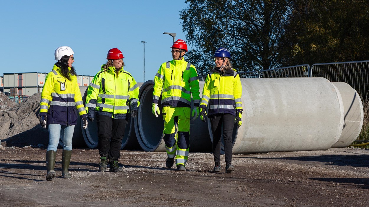Fyra personer i varselkläder som går mot kameran i bakgrunden syns ett stort betongrör.