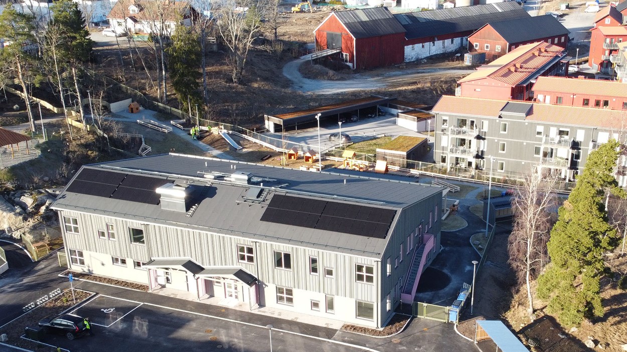 Drönarbild av förskolebyggnad med parkering framför, skogsdunge till vänster och bostadshus bakom och till höger.