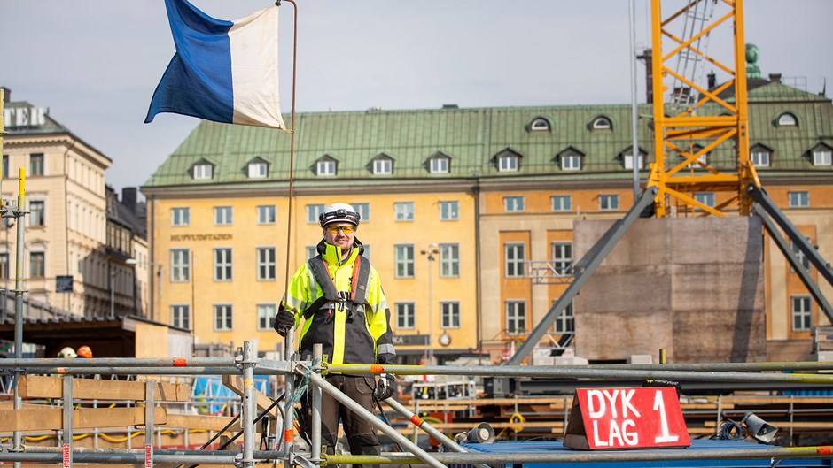 Martin Johansson står med full säkerhetsmundering vid den hissade blåvita dykarflaggan som visar att det är dykare i vattnet. På pråmen står det dyklag 1.