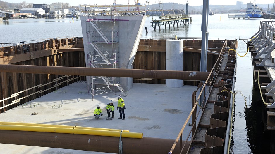 Tre personer men skyddskläder stå på betongfundament till Hisingsbron. I bakgrunden ser man hamnen och runtomkring är det vatten. 