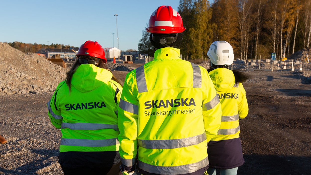 Tre kvinor sedda bakifrån i gula varselkläder där det står Skanskagymnasiet på en av ryggarna.