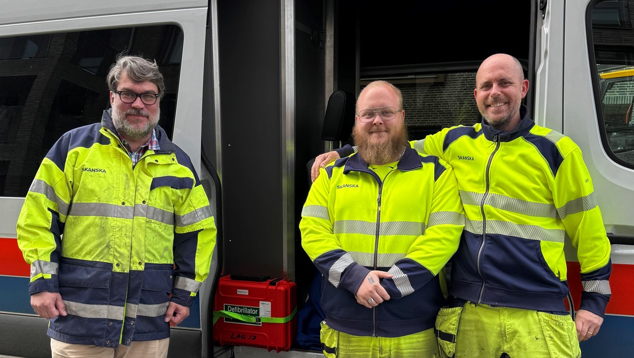 Tre män i varselkläder står framför en arbetsbil som utrustats med hjärtstartare.