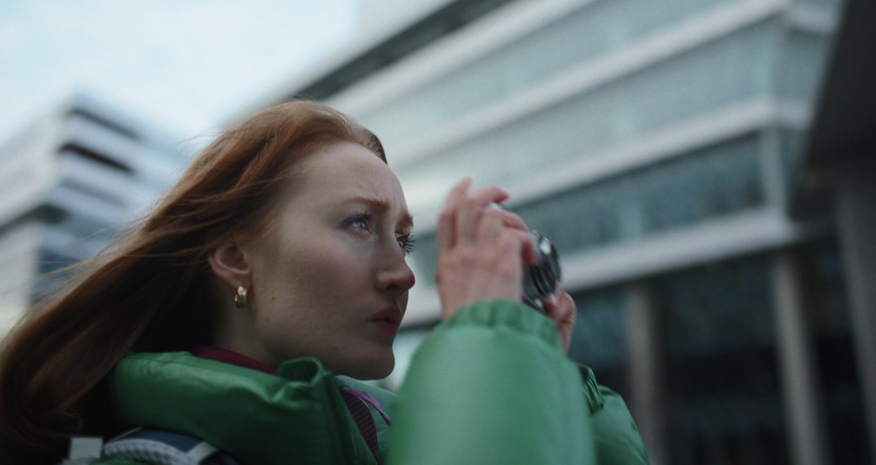 En kvinna i närbild med grön jacka som tittar åt höger och håller i en kamera framför ansiktet.