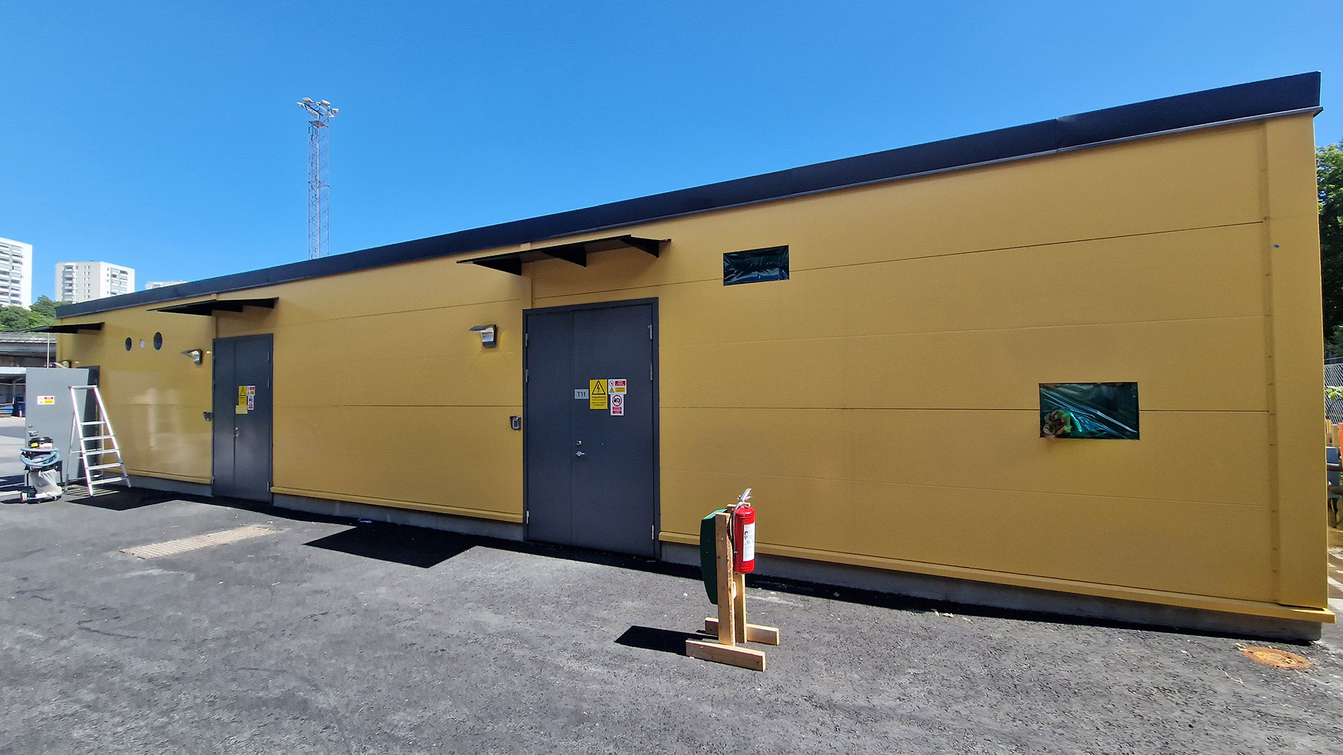 Gult teknikhus med dörr och ventilationsdetaljer, omgiven av ny asfalt. 