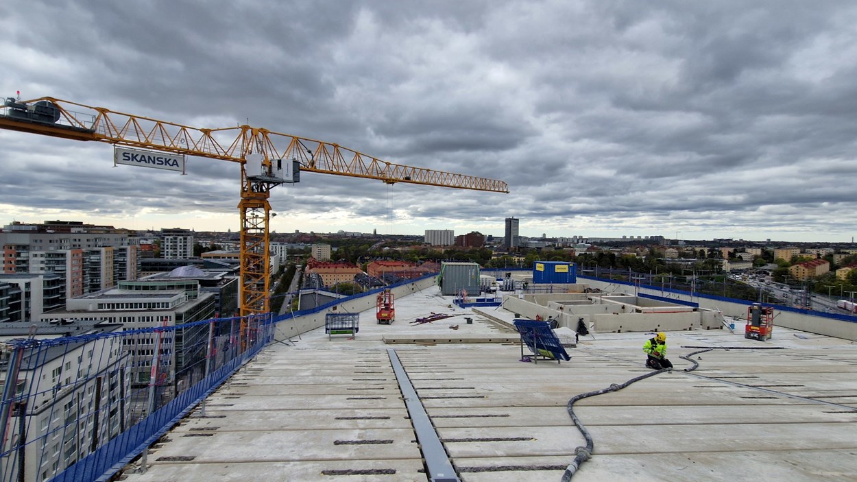 Byggarbetare på Olivins tak under byggnation, med Skanskas byggkran och vy över Stockholm i bakgrunden.