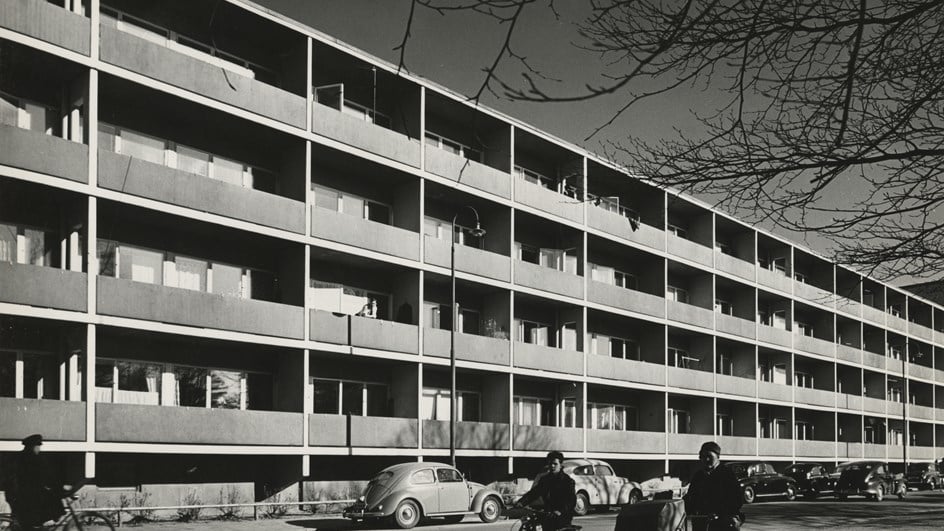 Svartvitt foto av flerbostadshus från 50-talet med "Folkabubblor" parkerade framför samt cyklister och en kvinna med barnvagn.