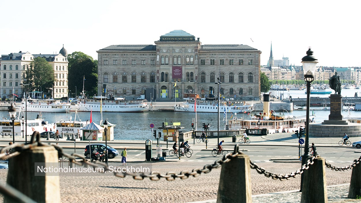 Pampig stenbyggnad längst bak i bilden. Vid kajen ligger vita passagerarbåtar och på kajen går ocoh cyklar människor.