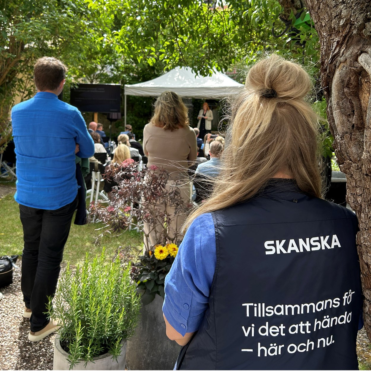 Personer som sitter och står i en trädgård och lyssnar till en kvinna som står på en scen och pratar.