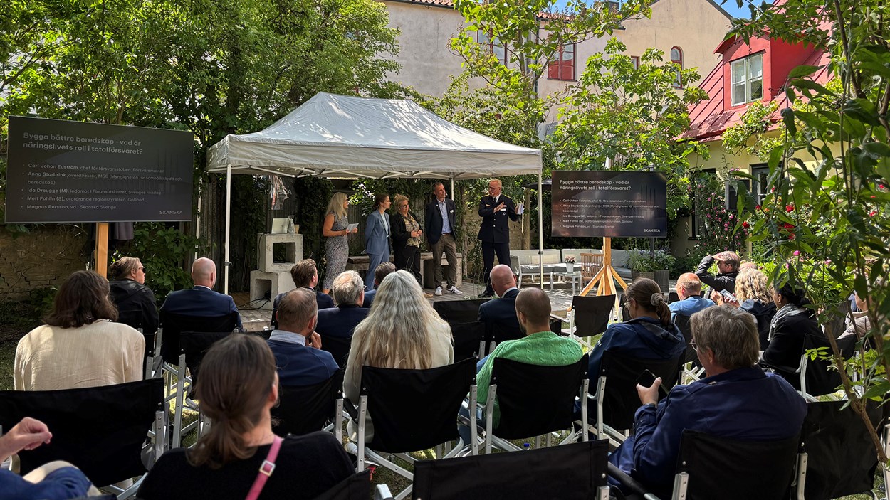 Besökare som sitter på stolar framför en utomhusscen i en grönskande trädgård. På scenen syns ett antal person er.