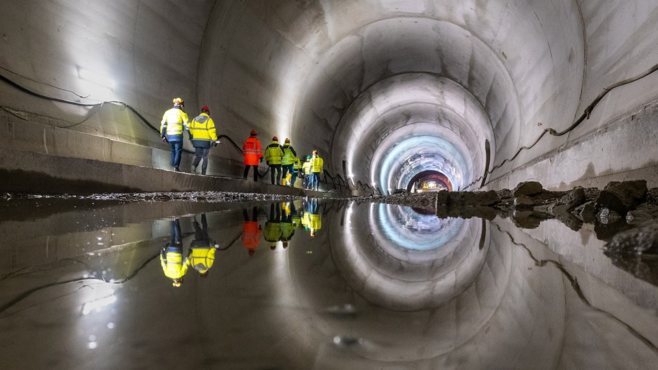 Interiör av en tunnel där ljuset i taket speglar sig i vattnet. På sidan av tunneln går personer som jobbar med tunnelbygget.