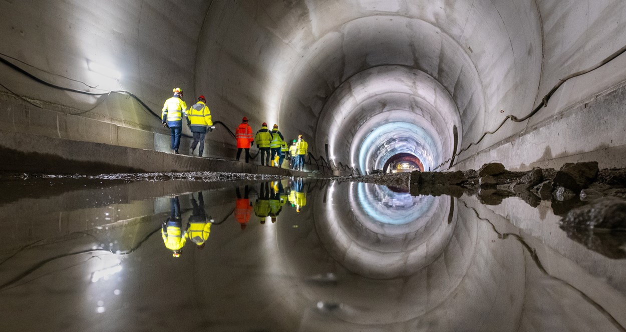 Interiör av en tunnel där ljuset i taket speglar sig i vattnet. På sidan av tunneln går personer som jobbar med tunnelbygget.