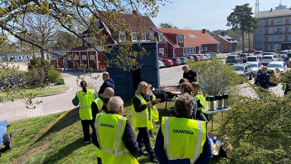 Elever, lärare och personal från Skanska grillar och äter korv i solen på en gräsplätt intill en parkering och en stor röd träbyggnad.