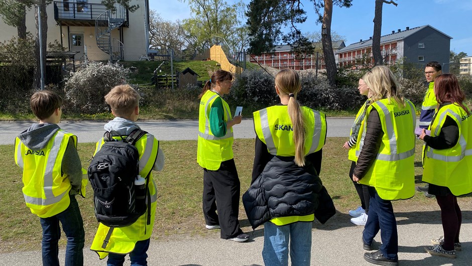 Skolelever och lärare i gula varselvästal lyssnar till personal från Skanska som pratar framför ett flerbostadshus.