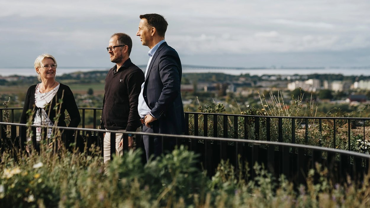 En kvinna och tvä män står på en öppen terrass högt upp. Långt bort i horisonten syns havet och en lång bro.