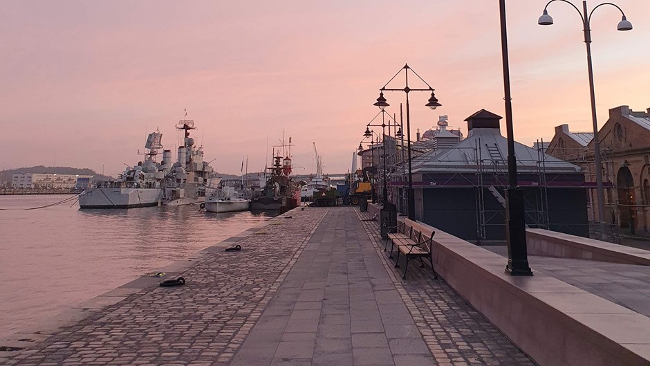 Kaj med mindre byggnader och intilliggande båtar i rosa morgonljus.