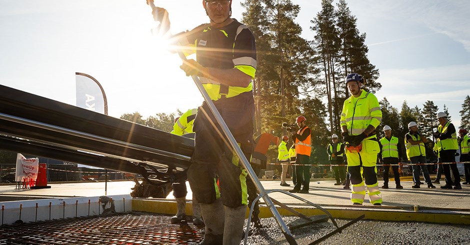 En man i blå och gula Skanska-kläder jämnar till betong på ett armeringsnät. I bakgrunden står kollegor och besökare.