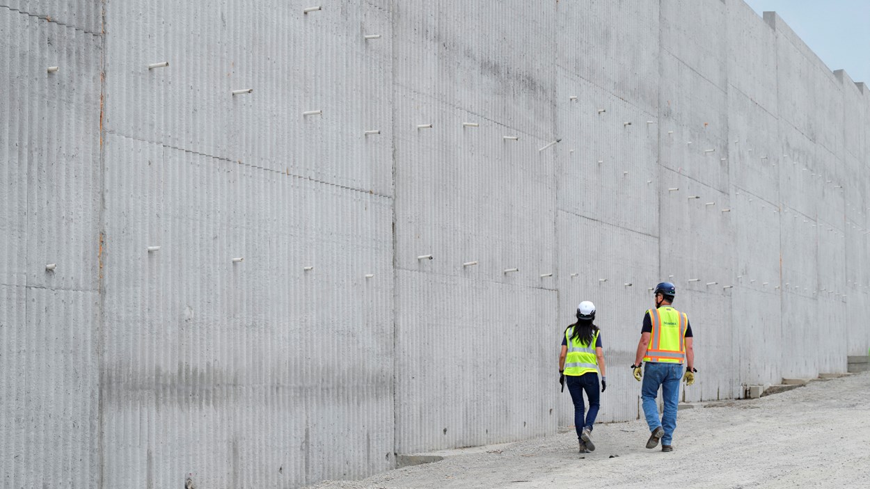 Två byggarbetare i varselvästar går utmed en hög betongvägg.