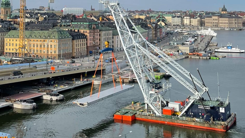 Lyftkran på vattnet lyfter och transporterar en gigantisk lucka i stål på 75 ton. I bakgrunden syns byggprojektet Slussen i Stockholm.