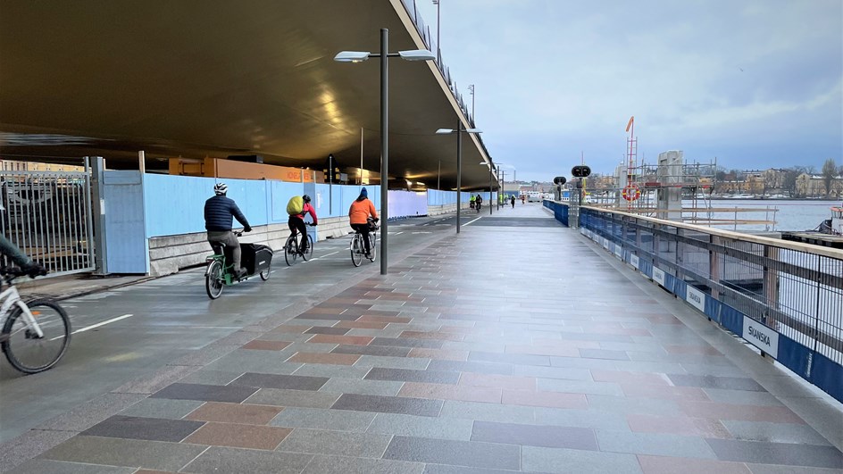 Cyklister på cykelbanan som går under den guldgula Slussenbron, till höger en plattbelagd gångväg utmed Saltsjön. I bakgrunden skymtar siluetten av Gamla stan.