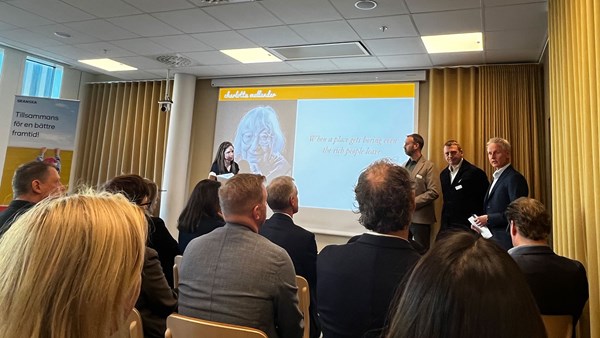 Människor sitter på rad i ett rum och lyssnar på en talare längst fram i lokalen.