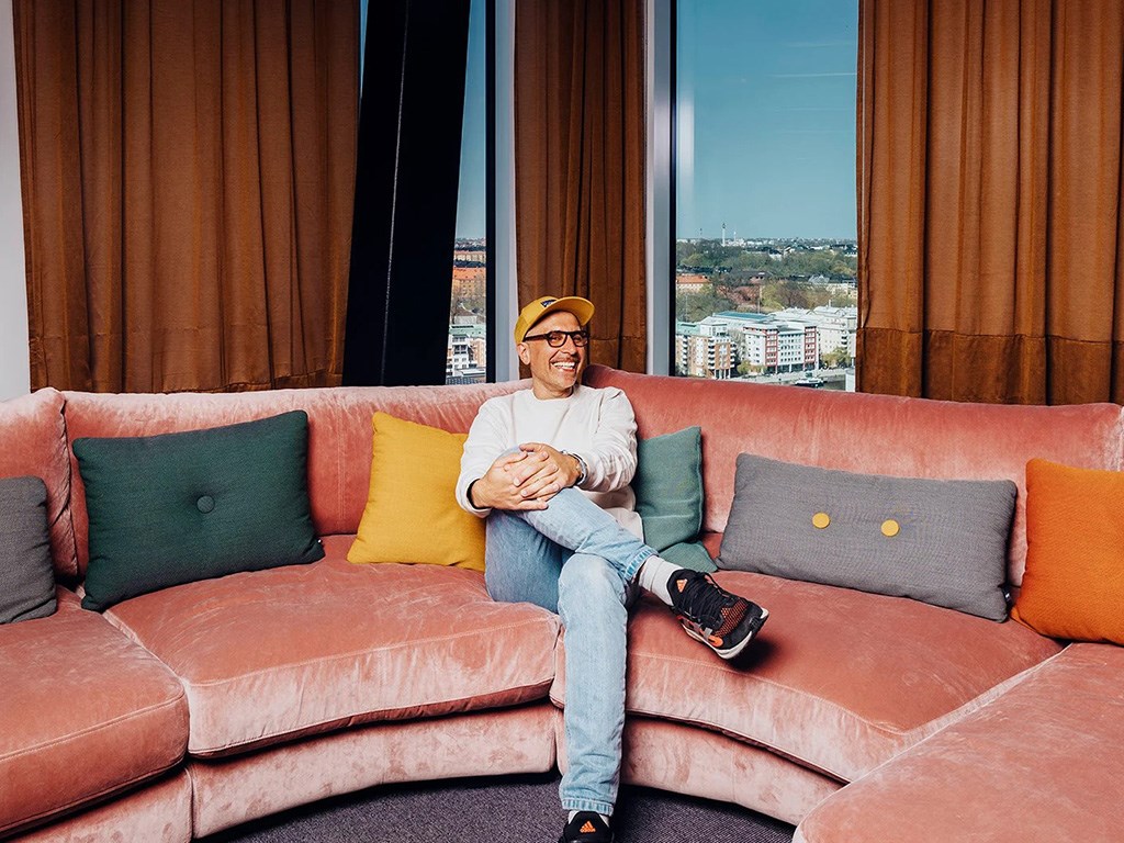 Man som skrattar och sitter i en soffa. I bakgrunden syns blå himmel aoch hustak.