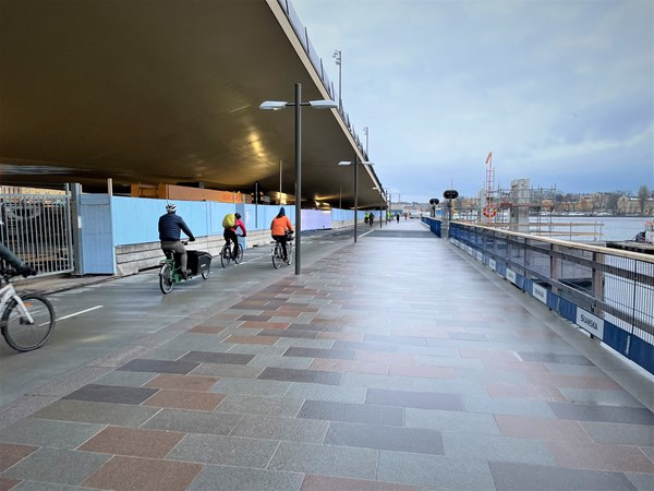 Cyklister på cykelbanan som går under den guldgula Slussenbron, till höger en plattbelagd gångväg utmed Saltsjön. I bakgrunden skymtar siluetten av Gamla stan.